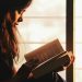 A woman engrossed in reading a book by a sunlit window creates a serene atmosphere.