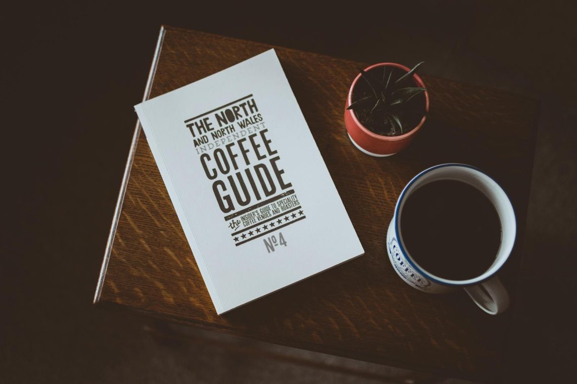 A cozy flat lay featuring a coffee guide, a mug of coffee, and a small plant on a wooden table.