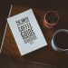 A cozy flat lay featuring a coffee guide, a mug of coffee, and a small plant on a wooden table.