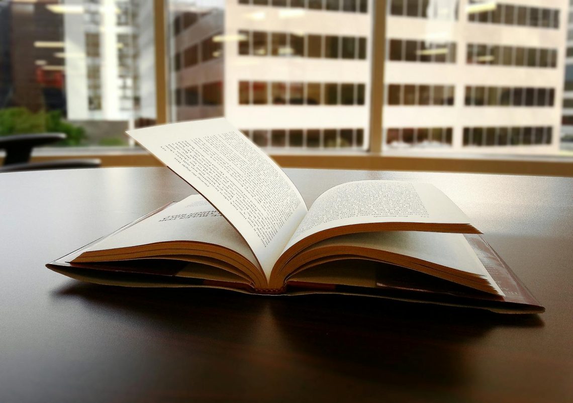 An open book sits on a desk in an office overlooking a city skyline through large windows.