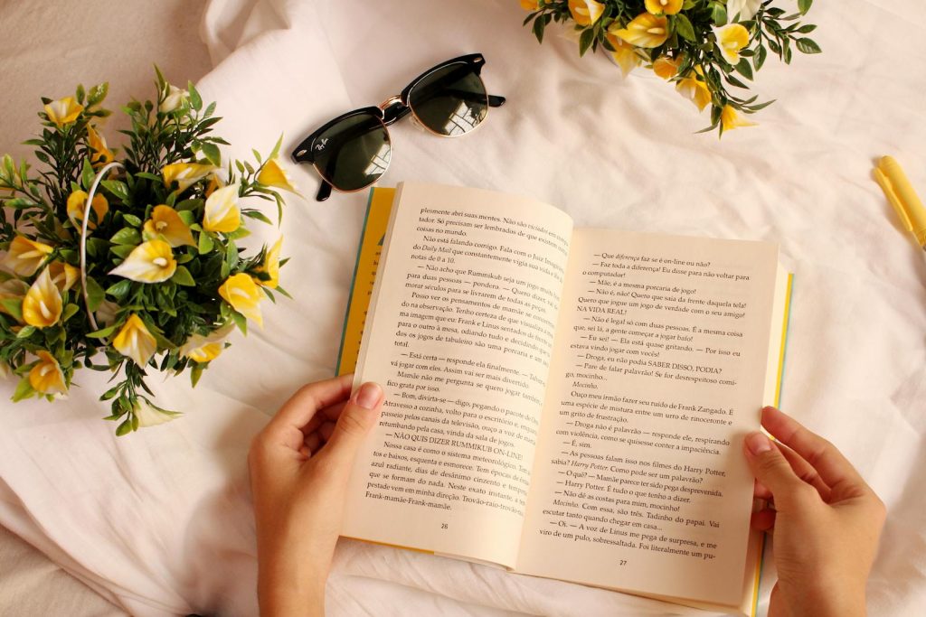 Hands holding a book surrounded by yellow flowers and sunglasses on a sunny day.