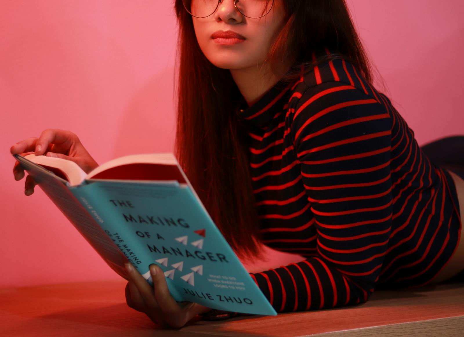 A woman lying on a pink background reading a book called 'The Making of a Manager'.