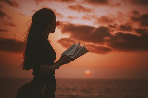 woman holding book while looking at body of water during golden hour