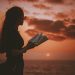 woman holding book while looking at body of water during golden hour